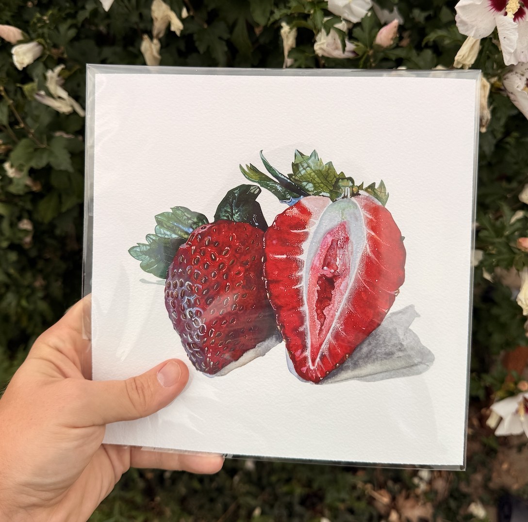 A hand holding a print of Strawberries, a watercolor painting by Wesley Ketcher, in front of a flowering shrub